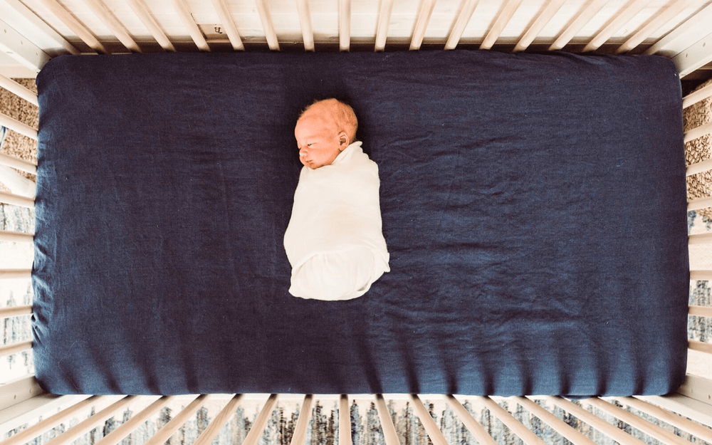 newborn baby in white blanket laying on navy blue linen crib sheet